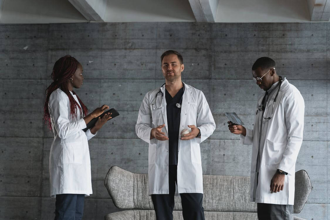 What You Need To Know About Outsourcing Medical Billing Three doctors standing wearing a white coat and stethoscope, one is holding a piece of paper, the second one is holding a baseball, and the third one is holding a tablet.